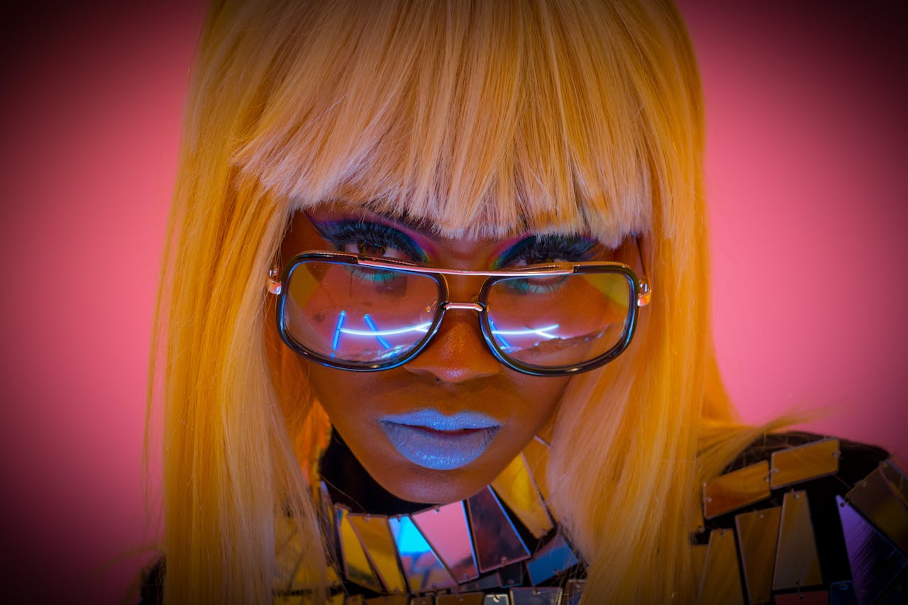 A bold and colorful portrait of a stylish woman wearing glasses and makeup against a pink background.